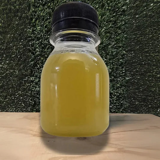 "Gember Shot Cold-Pressed Juice in een kleine doorzichtige fles met een goudgele kleur, gepresenteerd op een houten ondergrond tegen een groene achtergrond."