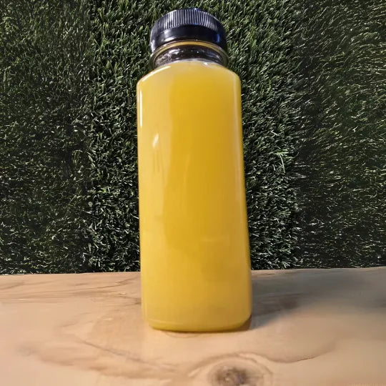 "Glow Cold-Pressed Juice in een doorzichtige fles met een felgele kleur, gepresenteerd op een houten ondergrond tegen een groene achtergrond."