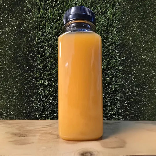 The Zest Cold-Pressed Juice in een doorzichtige fles met een oranje-gouden kleur, gepresenteerd op een houten ondergrond tegen een groene achtergrond.