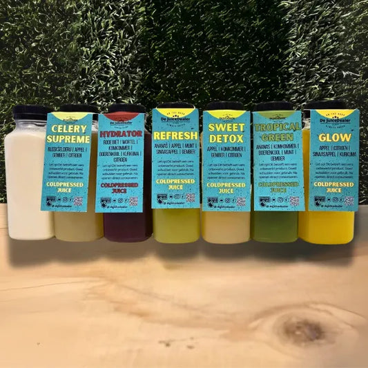 "Afbeelding van zes cold-pressed juices van De JuiceDealer met labels, op een houten oppervlak met een achtergrond van kunstgras. De juices zijn Celery Supreme, Hydrator, Refresh, Sweet Detox, Tropical Green en Glow."